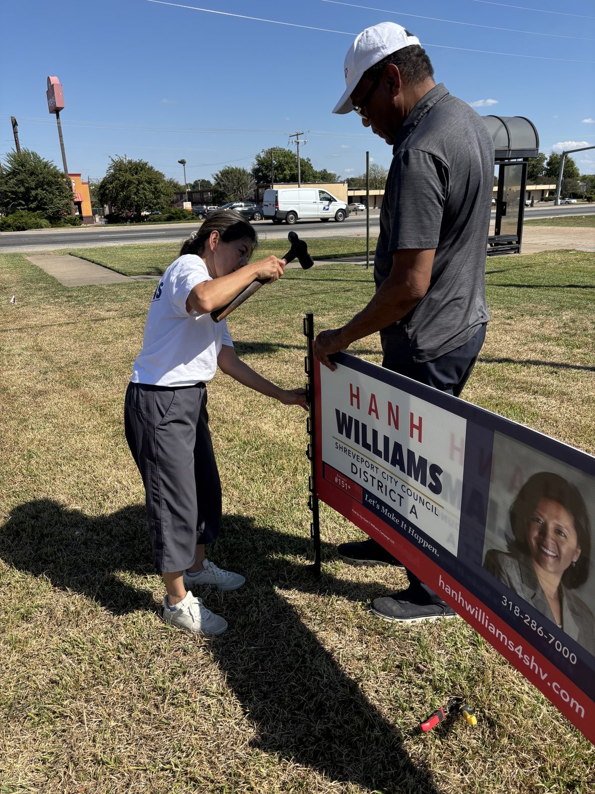 Hanh Williams with a hammer and a supporter putting a Hanh Williams City Council District A sign in the ground along a Shreveport street, October 7, 2026